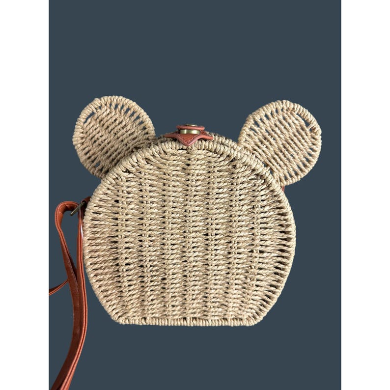 May include: A woven straw crossbody bag in the shape of a mouse, with two circular ears and a round body. The bag has a brown leather strap and a small leather accent on top. The bag is tan and brown.