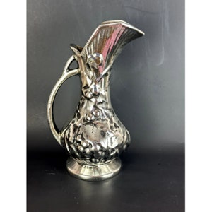 Puede incluir: Una jarra de plata con un diseño de vid. La jarra tiene una boca ancha y un asa.