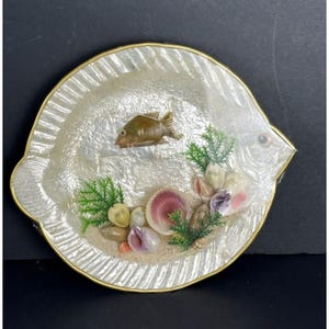 Op de afbeelding: Een decoratieve visvormige schaal met een parelmoer afwerking en een goudkleurige rand. De schaal heeft een kleine bruine vis, schelpen, groene planten en zand. Een uniek woondecoratie-item.