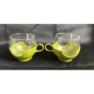 Puede incluir: Dos tazas de vidrio verde con un diseño único. Las tazas tienen un tazón de vidrio transparente y una base de plástico verde con un asa.