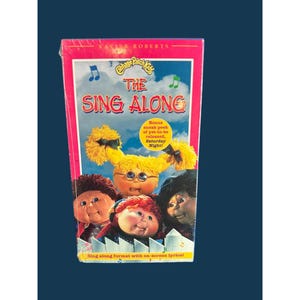 May include: A Cabbage Patch Kids The Sing Along VHS tape featuring Xavier Roberts branding. The cover displays four dolls with yarn hair against a blue sky background, with text including Bonus sneak peek of yet-to-be released, Saturday Night!