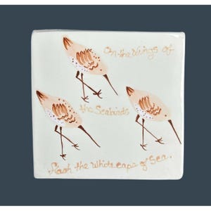 May include: Square ceramic tile with three stylized seabirds in beige and brown. The tile has handwritten text: "on the wings of the Seabirds, flash the white caps of Sea."