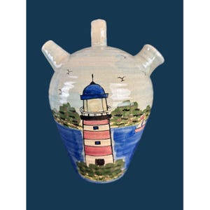 May include: A ceramic vessel with a rounded body, a central handle, and two side spouts. The surface features a hand-painted scene of a red and white striped lighthouse with a blue roof, set against a blue sea, green land, and small birds.