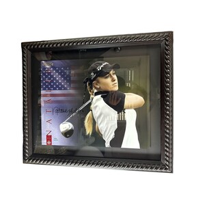 Puede incluir: Una foto enmarcada de una golfista con una camisa blanca y una falda negra, balanceando un palo de golf. La foto está firmada "Natalie Gulbis" y tiene una pequeña bandera estadounidense en el fondo. El marco es de color marrón oscuro con un diseño de cuerda.