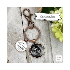 May include: A copper-toned keychain with a round charm featuring a black and white ultrasound image. The charm has the text "I can't wait to meet you Grandma!" and "Baby Maxwell December 2017".