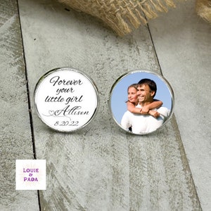 May include: Two round, silver-toned cufflinks. One cufflink features the text "Forever your little girl Allison 8.20.22" in black script. The other cufflink displays a color photo of a father and daughter. The cufflinks are on a gray surface.
