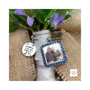 May include: A silver bridal bouquet charm with a blue rhinestone frame holding a photo of an older couple. The charm also has a round silver charm with the text "Everything I Am, you Helped me To Be".