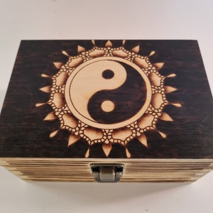 May include: A wooden box with a hinged lid and a latch closure. The top of the box is decorated with a black and white yin yang symbol surrounded by a mandala design. The box is stained a dark brown colour.