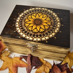 May include: A wooden box with a dark brown stain and a sunflower mandala design burned into the lid. The box has a metal latch and is surrounded by fall leaves.