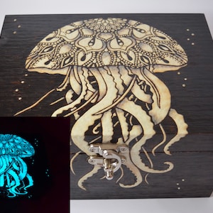 May include: A wooden box with a glow-in-the-dark jellyfish design. The jellyfish is intricately detailed with a mandala pattern on its bell and flowing tentacles. The box has a silver latch and is made of dark wood.