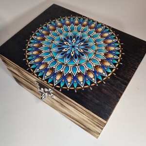 Può includere: Una scatola di legno con un design a mandala blu, bianco e marrone sul coperchio. La scatola ha una chiusura argentata.