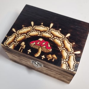 Può includere: Una scatola di legno con un coperchio incernierato e una chiusura. La scatola è decorata con un disegno pirografato di un fungo con cappello rosso e un motivo a mandala.