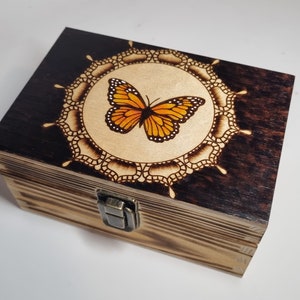 Può includere: Una scatola di legno con una macchia marrone scuro e una base in legno chiaro. Il coperchio presenta un disegno di farfalla bruciato a legno all'interno di un mandala. La scatola ha una chiusura in ottone.