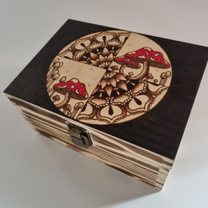 Può includere: Una scatola di legno con un coperchio incernierato e una chiusura. Il coperchio è decorato con un motivo a mandala bianco e nero con tre funghi rossi.
