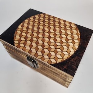 Puede incluir: Una caja de madera con una mancha marrón oscura y un patrón geométrico grabado con láser en la tapa. La caja tiene un cierre de metal.