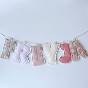 May include: A fabric letter banner spelling the name FREYJA, featuring individual stuffed letters in various patterns and colours including floral prints, solid light pink, polka dots, and cream, all suspended from a white twisted cord.