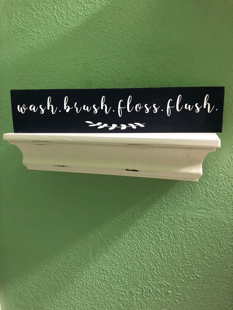 May include: Black wooden sign with white text that reads "wash.brush.floss.flush."  The sign is sitting on a white shelf.