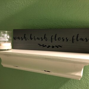 May include: A gray wooden sign with the text "wash. brush. floss. flush." in black lettering. A small white candle is on a white shelf below the sign.