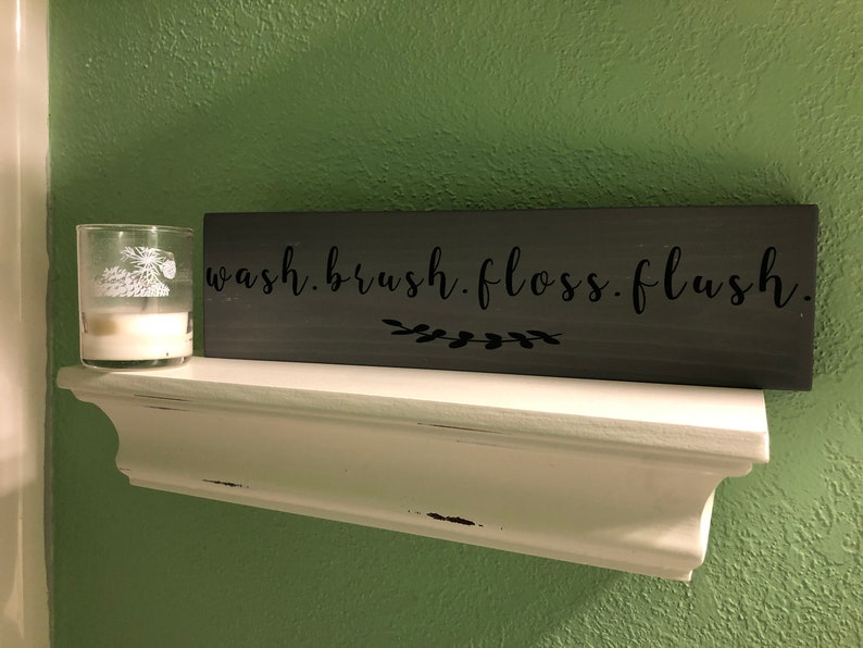 May include: A gray wooden sign with the text "wash. brush. floss. flush." in black lettering. A small white candle is on a white shelf below the sign.