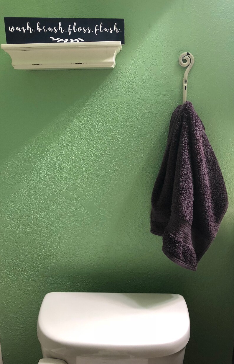 May include: A white shelf with the words "wash. brush. floss. flush." on a black sign. A dark gray towel hangs on a decorative hook on a light green wall. A white toilet tank is in the lower right corner of the image.