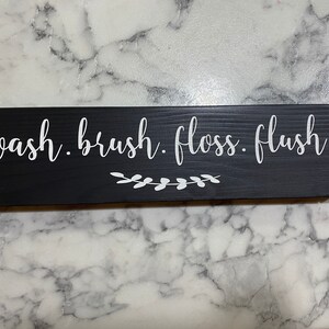 May include: Black wooden sign with white text that reads "wash. brush. floss. flush." with a small white floral design underneath.
