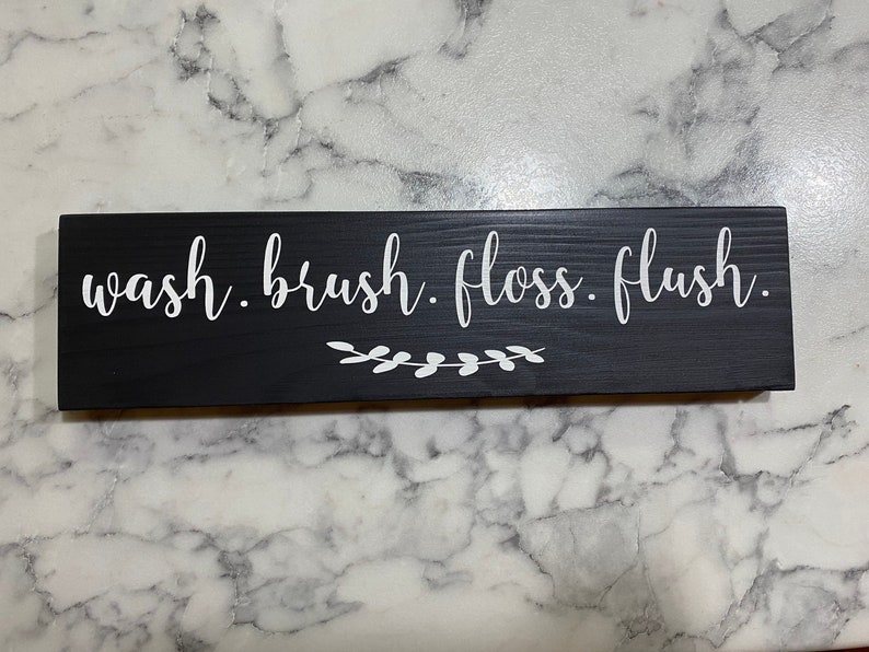 May include: Black wooden sign with white text that reads "wash. brush. floss. flush." with a small white floral design underneath.