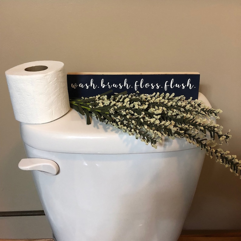 May include: A white toilet with a roll of toilet paper and a blue wooden sign that says "wash. brush. floss. flush." on top of it. There is a sprig of white flowers on the toilet tank.