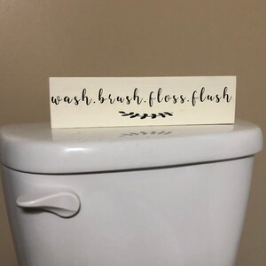 May include: White wooden sign with black text that reads "wash. brush. floss. flush" with a small black floral design below the text. The sign is sitting on top of a white toilet.