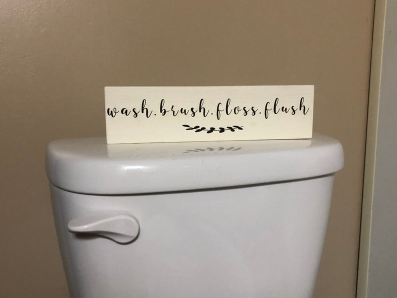 May include: White wooden sign with black text that reads "wash. brush. floss. flush" with a small black floral design below the text. The sign is sitting on top of a white toilet.