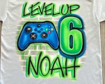 May include: White t-shirt with a green and blue airbrushed design. The design features a video game controller, the number 6, and the name "Noah". The text "Level Up" is also included in the design.