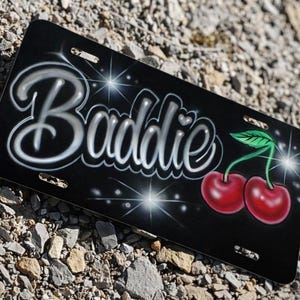 May include: Black license plate with the word "Baddie" in a stylized, airbrushed font. Features two red cherries with green leaves. The background has a sparkling effect.