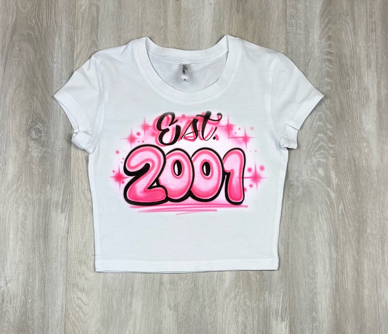 May include: White crop top with the text "Est. 2001" in a bubble-style font. The text is outlined in black and filled with pink, with pink starburst accents.
