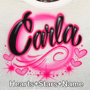 May include: White t-shirt with the name "Carla" in black, outlined in pink, within a pink airbrushed circle. Pink hearts and starbursts surround the name. The text "Hearts+Stars+Name" is at the bottom.