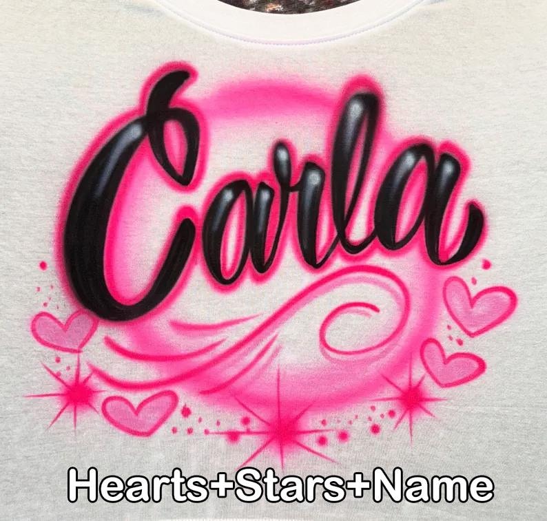 May include: White t-shirt with the name "Carla" in black, outlined in pink, within a pink airbrushed circle. Pink hearts and starbursts surround the name. The text "Hearts+Stars+Name" is at the bottom.