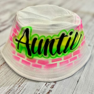 May include: White bucket hat with a pink and green brick pattern and the word "Auntie" written in black.