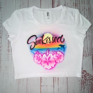 May include: White crop top with a round neckline. The shirt features a colorful graphic with the text "Sun Kissed", a sunset, dolphins, and pink hibiscus flowers. The shirt is a medium size.