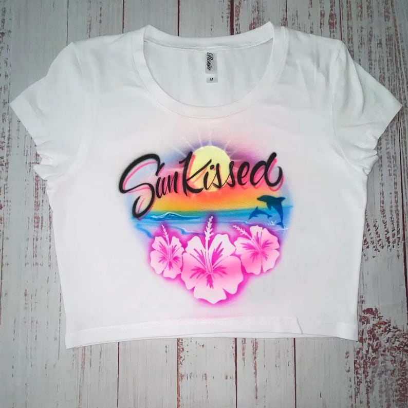 May include: White crop top with a round neckline. The shirt features a colorful graphic with the text "Sun Kissed", a sunset, dolphins, and pink hibiscus flowers. The shirt is a medium size.