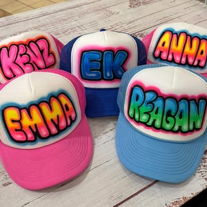 May include: Five colorful trucker hats with personalized names. Each hat features a white front panel with a name in a bubble letter style, outlined in a contrasting color. The hats have mesh backs and curved brims in various colors, including pink and blue.