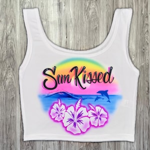 May include: White crop top with a colorful airbrushed design. The design includes the words "Sun Kissed" in black script, a sunset, ocean waves, two dolphins, and hibiscus flowers. The top has a scoop neck and sleeveless design.