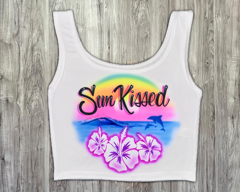 May include: White crop top with a colorful airbrushed design. The design includes the words "Sun Kissed" in black script, a sunset, ocean waves, two dolphins, and hibiscus flowers. The top has a scoop neck and sleeveless design.