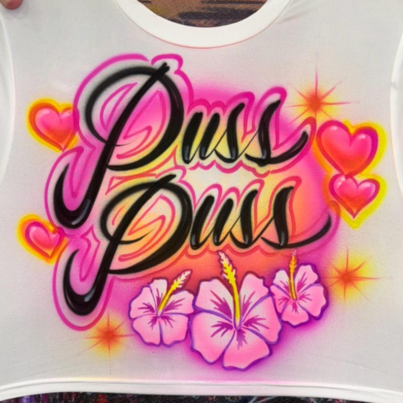 May include: White crop top with the text "Puss Puss" in a black, pink, and yellow airbrushed design. The design includes pink hearts, yellow stars, and pink hibiscus flowers.
