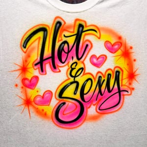 May include: White t-shirt with an airbrushed design. The design features the words "Hot & Sexy" in black script outlined in yellow and pink. Pink hearts and orange starbursts surround the text.