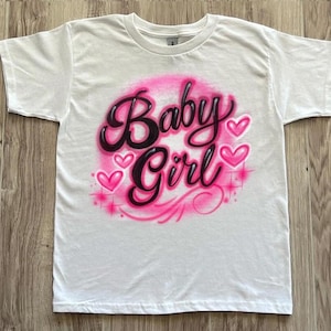 May include: White t-shirt featuring the words "Baby Girl" in a pink and black airbrushed design. The design includes pink hearts and starbursts, creating a playful and feminine aesthetic.
