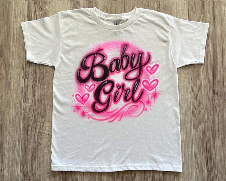 May include: White t-shirt featuring the words "Baby Girl" in a pink and black airbrushed design. The design includes pink hearts and starbursts, creating a playful and feminine aesthetic.