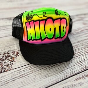 May include: A black mesh trucker hat with a rainbow gradient design featuring the name "NIKOTB" in large, colorful letters. The hat also has a small musical note design.