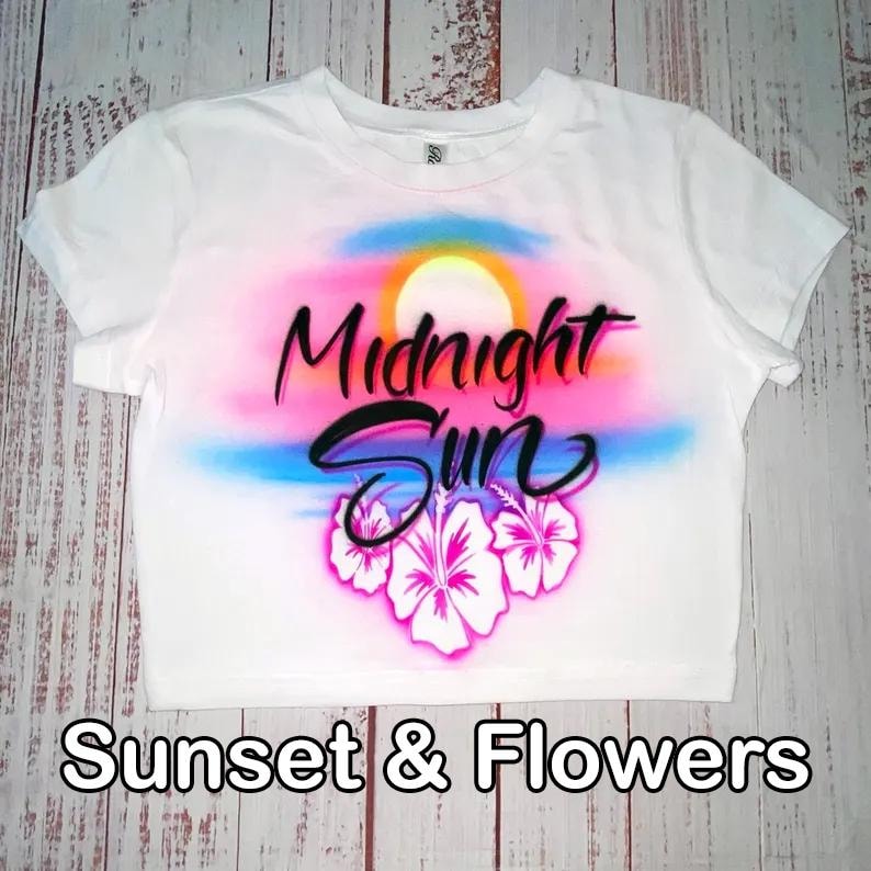 May include: White crop top with a colorful airbrushed design featuring a sunset, hibiscus flowers, and the words "Midnight Sun". The text "Sunset & Flowers" is printed at the bottom of the shirt.