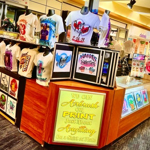 May include: A shop display featuring custom airbrushed t-shirts with diverse designs, including portraits and text. The shirts are displayed on mannequins. A sign reads, "We Can Airbrush or Print Just About Anything On a Shirt or Plate."