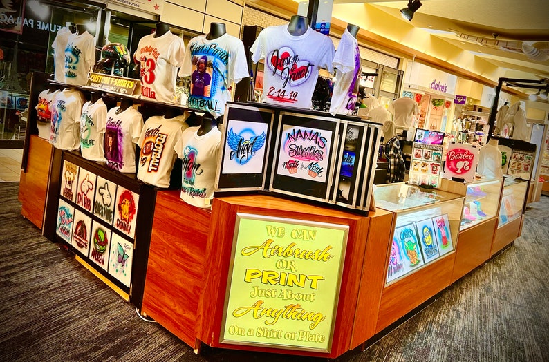 May include: A shop display featuring custom airbrushed t-shirts with diverse designs, including portraits and text. The shirts are displayed on mannequins. A sign reads, "We Can Airbrush or Print Just About Anything On a Shirt or Plate."