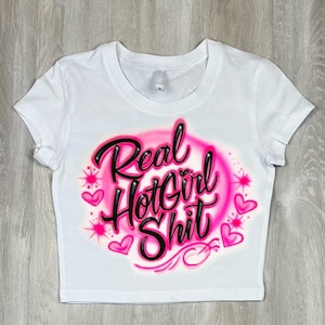 May include: White crop top with the text "Real Hot Girl Shit" in black script outlined in pink, surrounded by a pink airbrushed design with hearts and starbursts. The shirt has short sleeves and a crew neck.