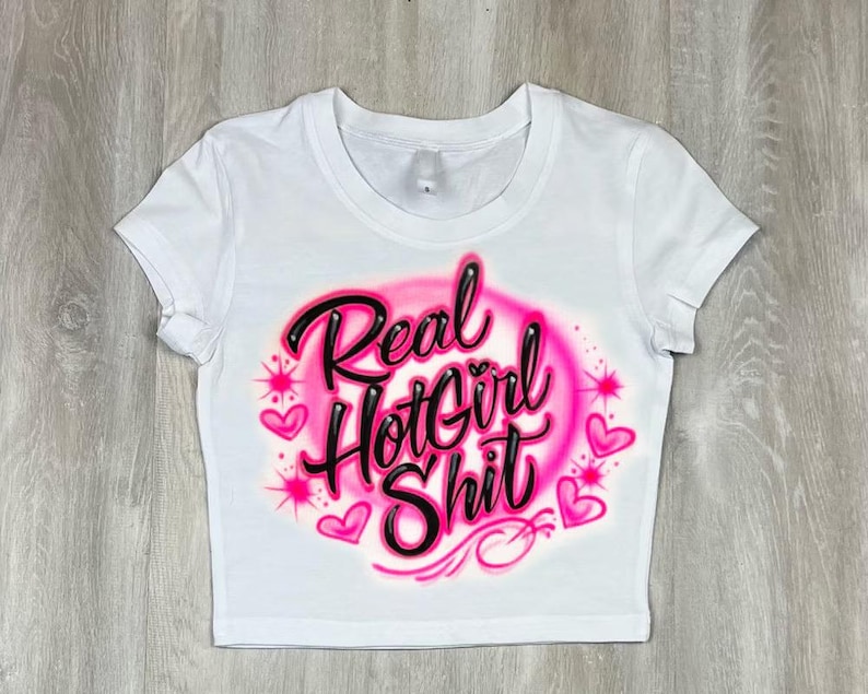 May include: White crop top with the text "Real Hot Girl Shit" in black script outlined in pink, surrounded by a pink airbrushed design with hearts and starbursts. The shirt has short sleeves and a crew neck.
